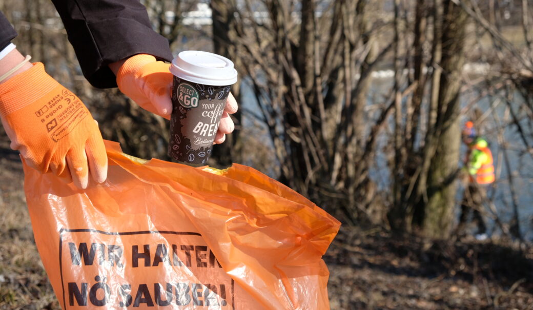 Auf dem Bild ist eine Person zu sehen, die mit orangen Handschuhen einen Coffee-to-go-Becher aufnimmt und in einen orangenen Müllsack wirft. Im Hintergrund sind weitere Personen in Warnwesten an einem Flussufer beschäftigt, offenbar bei einer Fluss- oder Uferreinigungsaktion. Auf dem Müllsack steht: „WIR HALTEN NÖ SAUBER!“.