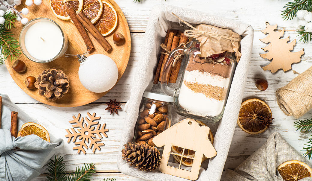 In einem Glas verpackte Nüsse, Zimtstangen, getrocknete Orangenschalen und weitere Weihnachtsdeko und in Tücher verpackte Geschenke sind auf dem Bild zu sehen.