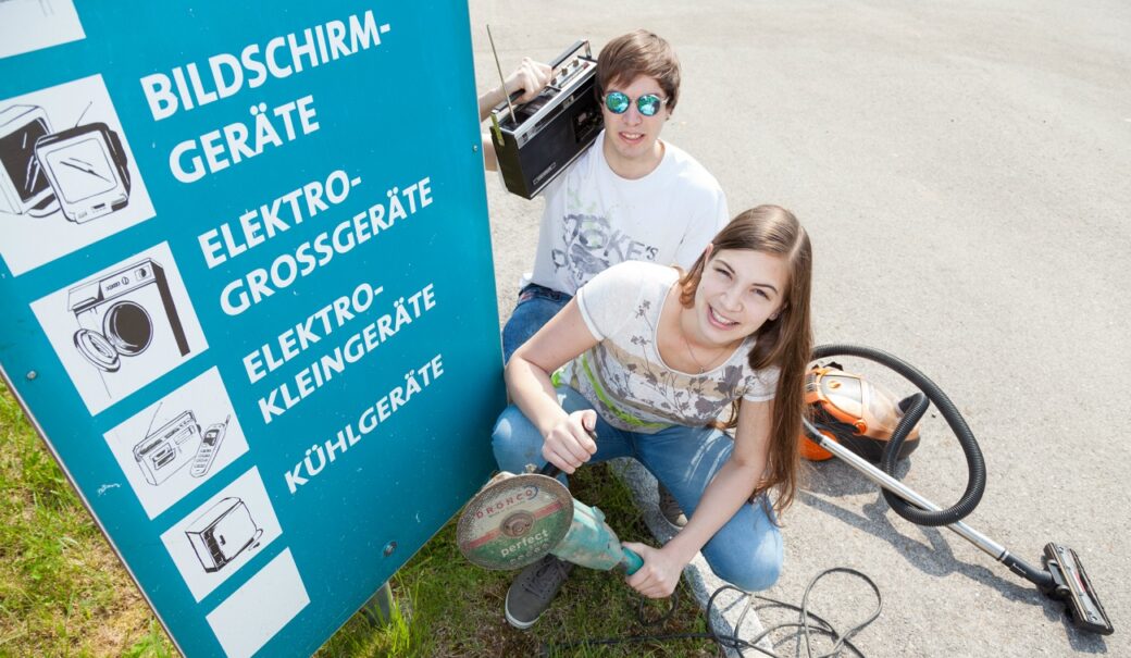 Zwei Jugendliche mit Elektroaltgeräte vor einer Trenn-Infotafel am Sammelzentrum